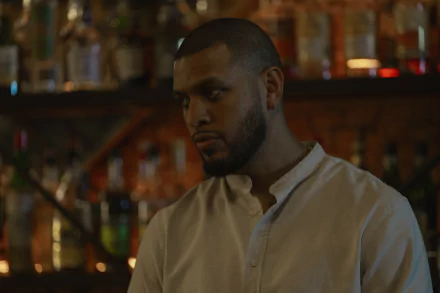 A HD desktop wallpaper featuring a scene from the TV Show Presumed Innocent (2024) with a man standing in a dimly-lit bar.