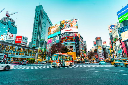 HD desktop wallpaper depicting a vibrant Tokyo city street with bustling traffic, towering buildings, and colorful advertisements. A bus is driving through a busy intersection, capturing the essence of urban life in Japan.