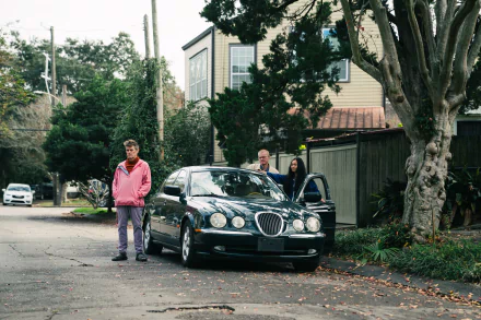 A scene from the movie Kinds of Kindness featuring three people with a luxury car on a residential street, framed by trees and houses. HD desktop wallpaper and background.