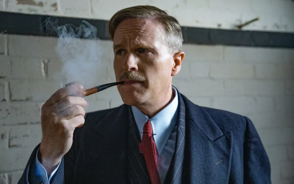 A man in a vintage suit and tie, with a neat haircut, smoking a pipe against a brick wall background, in a still from the movie The Ministry of Ungentlemanly Warfare. This image serves as a high-definition desktop wallpaper.