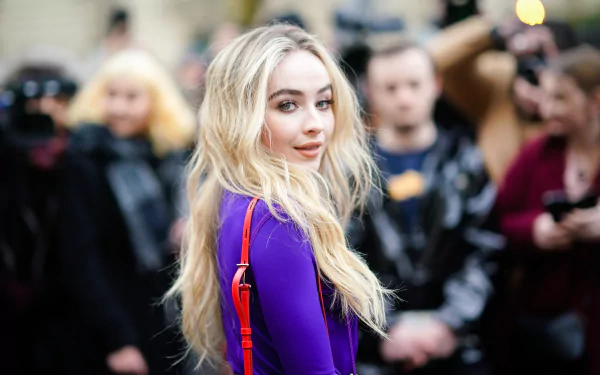 Blonde singer Sabrina Carpenter in a vibrant purple outfit, captured in sharp 4K Ultra HD as she walks past a blurred crowd.