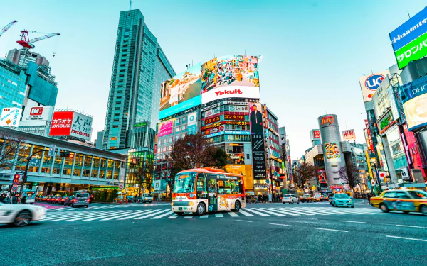 HD desktop wallpaper depicting a vibrant Tokyo city street with bustling traffic, towering buildings, and colorful advertisements. A bus is driving through a busy intersection, capturing the essence of urban life in Japan.