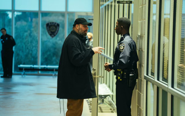 HD desktop wallpaper and background featuring a scene from the movie Kinds of Kindness, showing a man in a cap speaking with a uniformed officer in a well-lit indoor area.