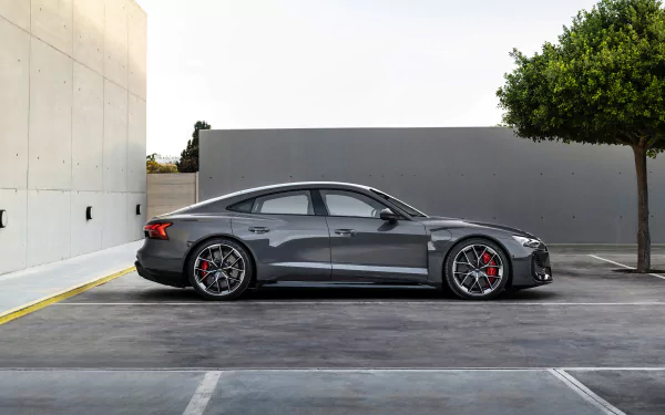 HD desktop wallpaper of an Audi RS e-Tron GT parked in a sleek, minimalist setting with modern architecture and a tree. The car's metallic finish and striking design are prominently displayed.