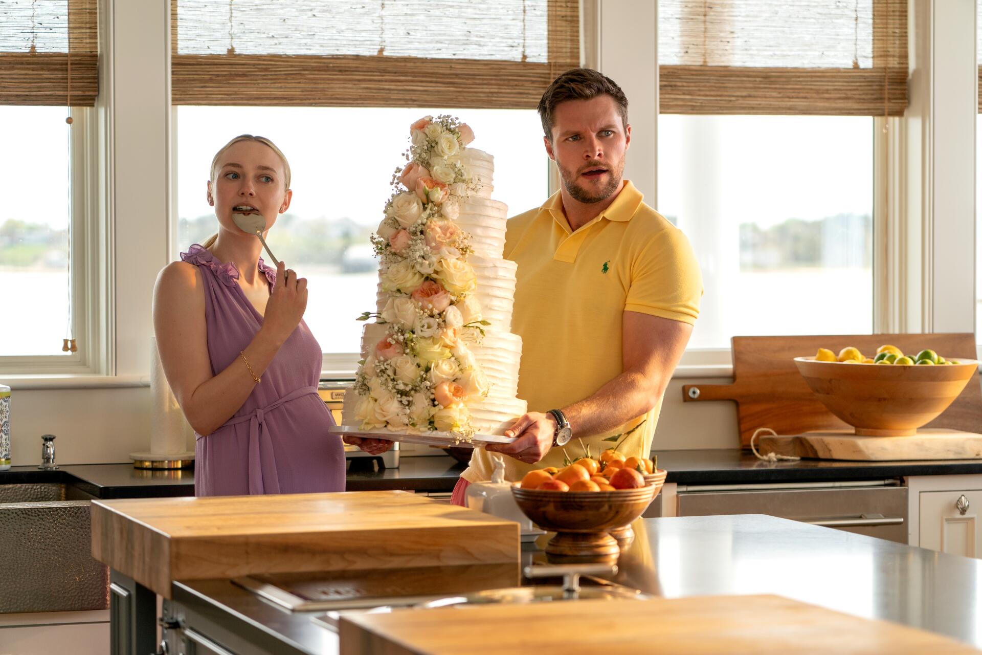 Dakota Fanning and Jack Reynor in a scene from the TV show The Perfect Couple, featuring a celebration with a large wedding cake in a bright kitchen setting.
