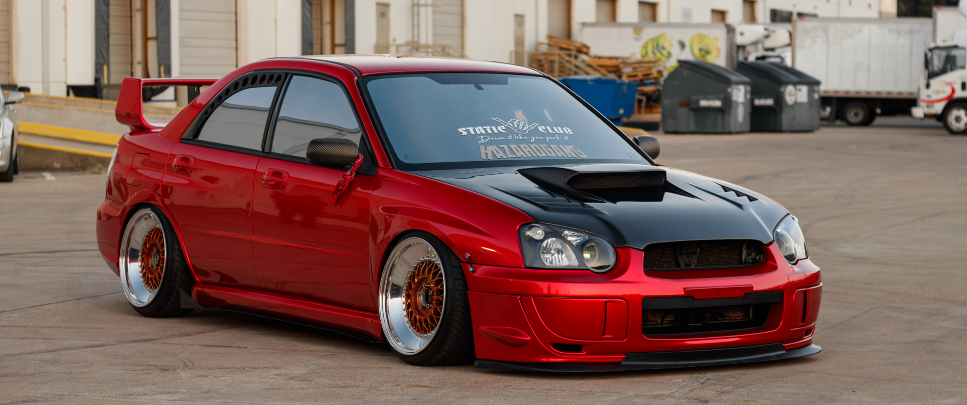 A striking red Subaru Impreza WRX STI, featuring a bold black hood and lowered stance, showcased in an HD wallpaper setting with an urban backdrop.