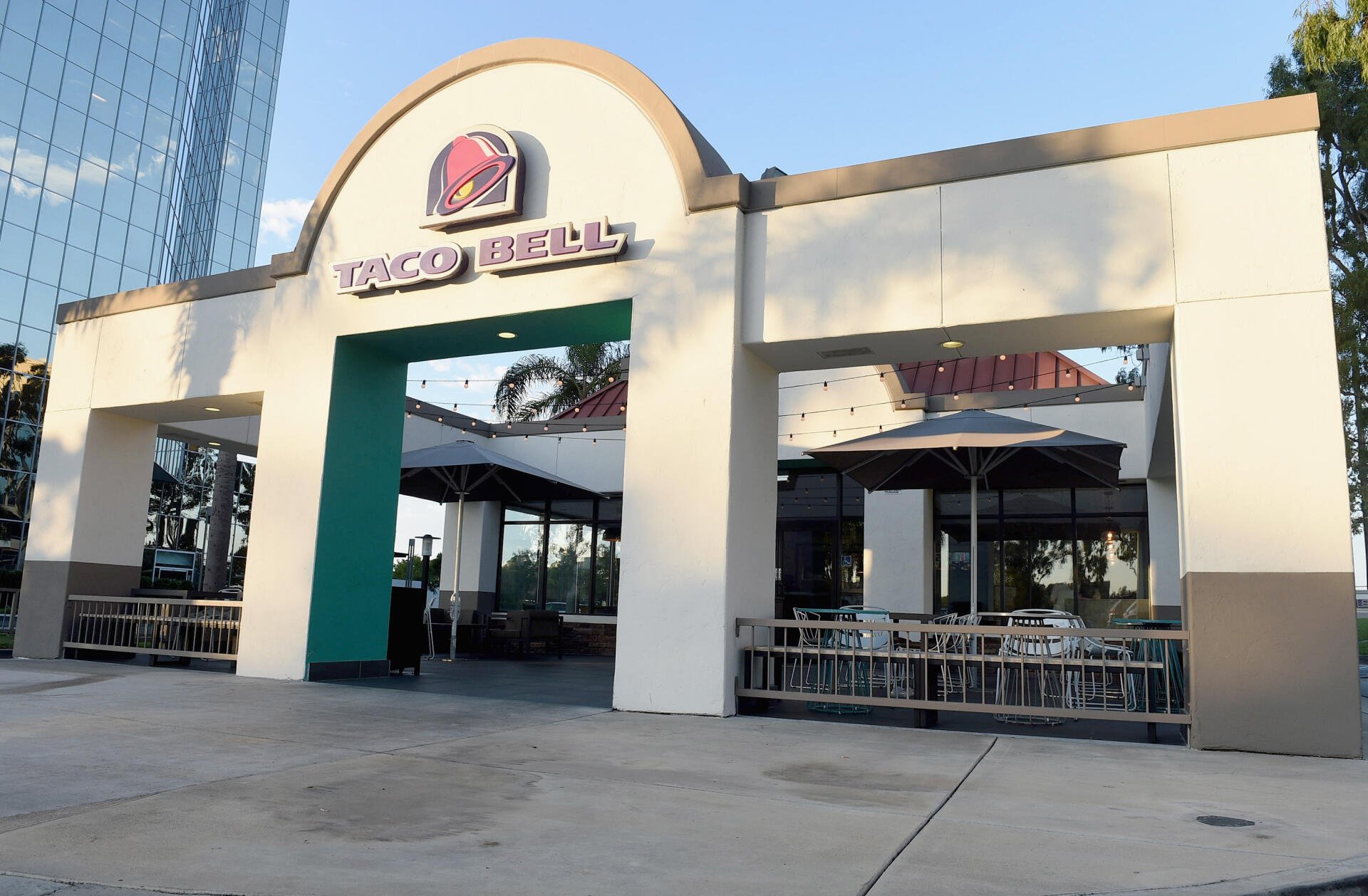 A high-definition desktop wallpaper showcasing a Taco Bell restaurant exterior, featuring modern architecture and outdoor seating under umbrellas.