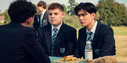 A scene from the TV show Heartstopper, featuring two characters in school uniforms at a table, engaged in conversation, with a bottle of water and food in the foreground.