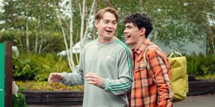 Two young men laugh and share a moment outdoors, surrounded by greenery, in a scene from the TV show Heartstopper. The image is a vibrant 4K Ultra HD wallpaper.