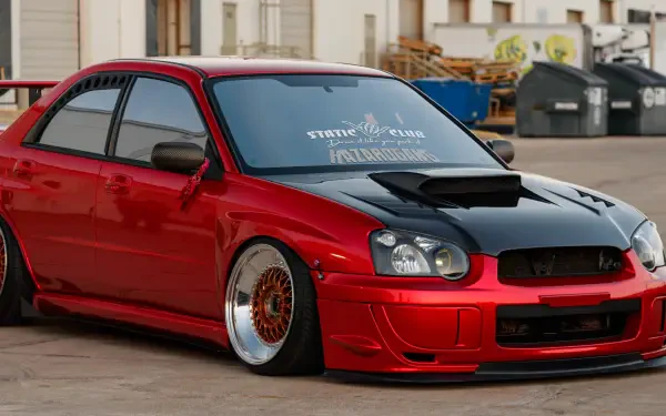 A striking red Subaru Impreza WRX STI, featuring a bold black hood and lowered stance, showcased in an HD wallpaper setting with an urban backdrop.