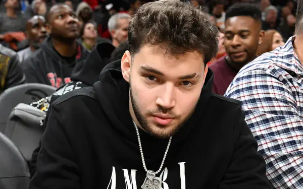 An HD PC desktop wallpaper featuring streamer Adin Ross, focused and seated among a crowd at a lively event, showcasing a casual style with a unique necklace.