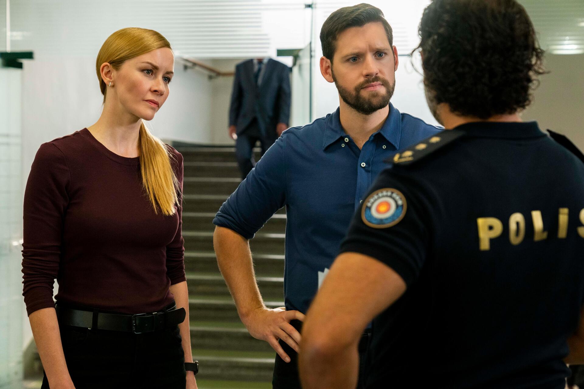 A scene from the TV show *FBI: International*, featuring two agents engaging in a serious conversation with a police officer. The backdrop includes a modern interior setting.