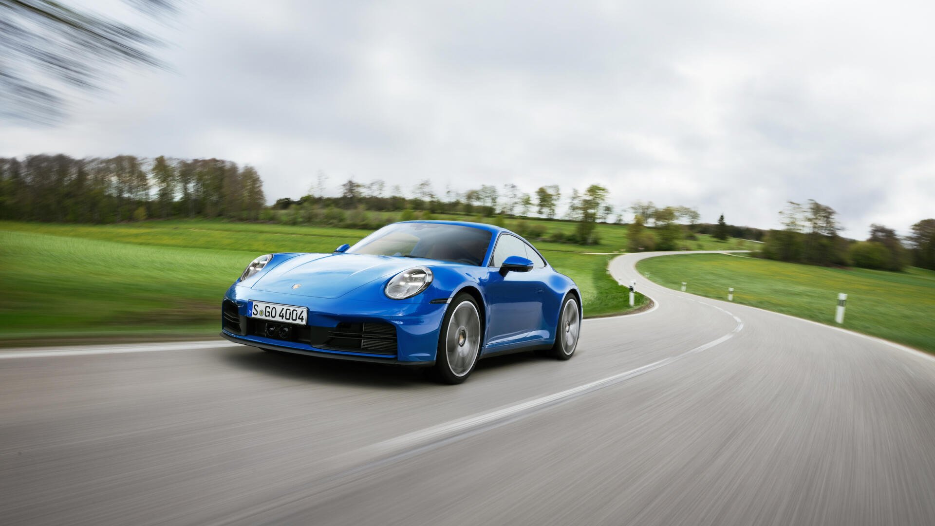 A sleek blue Porsche 911 Carrera speeds along a winding road, surrounded by lush greenery, showcasing its dynamic design and performance. An HD desktop wallpaper for car enthusiasts.