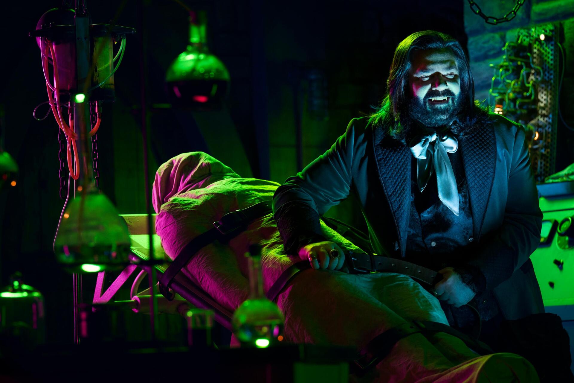 A vibrant HD wallpaper from What We Do in the Shadows, featuring Matt Berry in a dimly lit lab, animatedly interacting with a figure wrapped in a shroud, enhanced by colorful lighting effects.