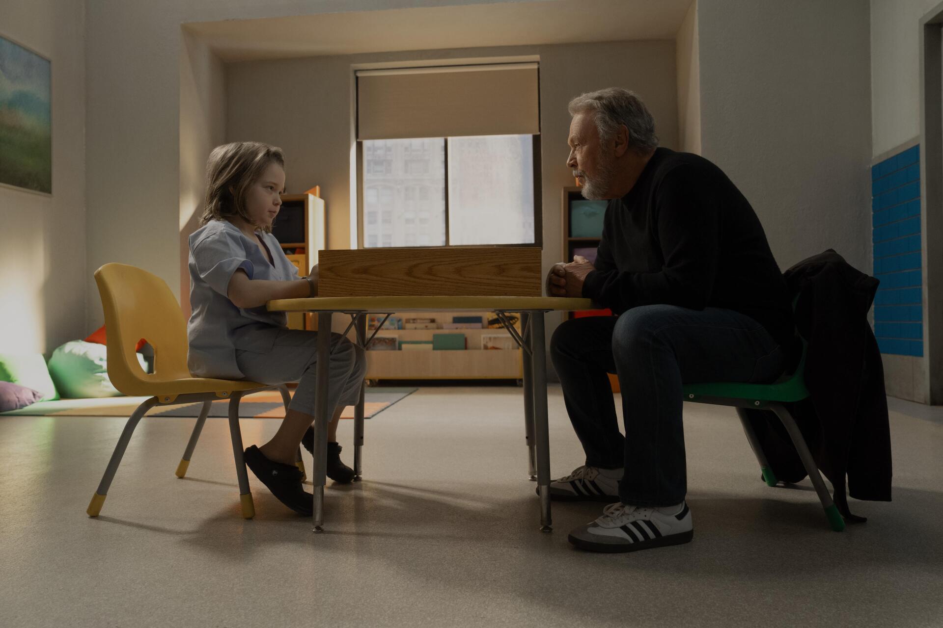 A scene from the TV show Before (2024) featuring Billy Crystal seated across from a young girl at a table, both engaged in a contemplative moment. The setting is bright and simple.