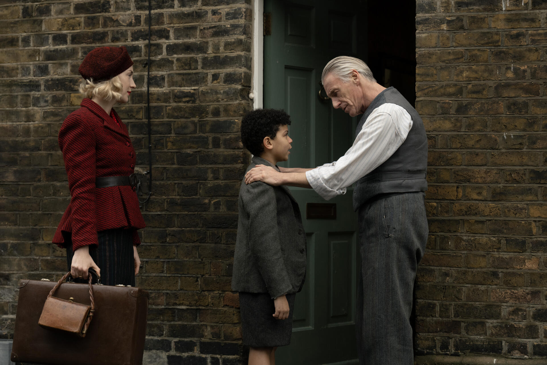A scene from the movie Blitz (2024), featuring a woman in a red coat, a boy in a suit, and an elderly man in a vest, set against a brick wall and door backdrop. 8K Ultra HD quality.