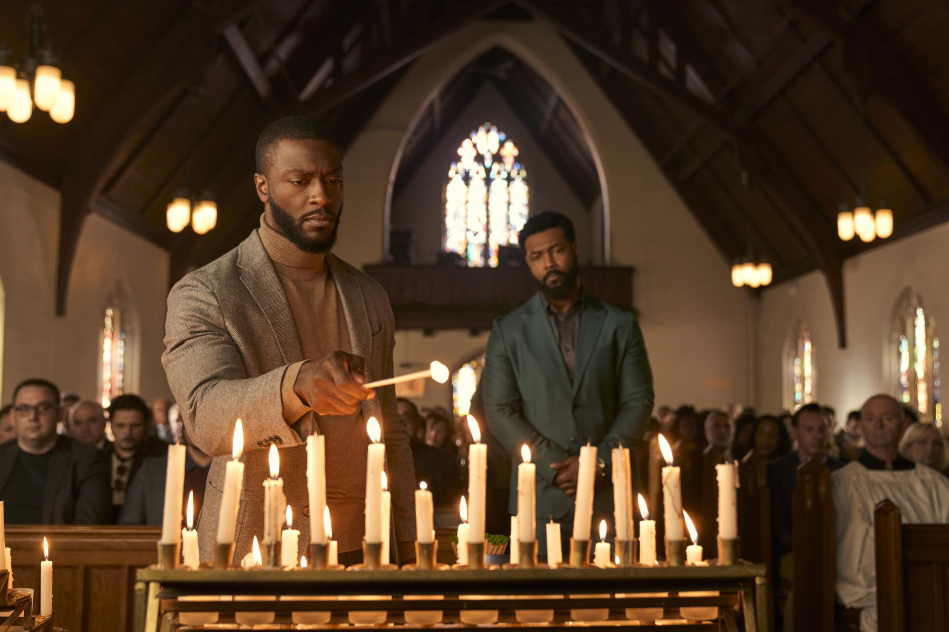 Aldis Hodge and Isaiah Mustafa in a poignant scene from the TV show Cross (2024), set in a candlelit church filled with attendees. An evocative backdrop for your desktop.