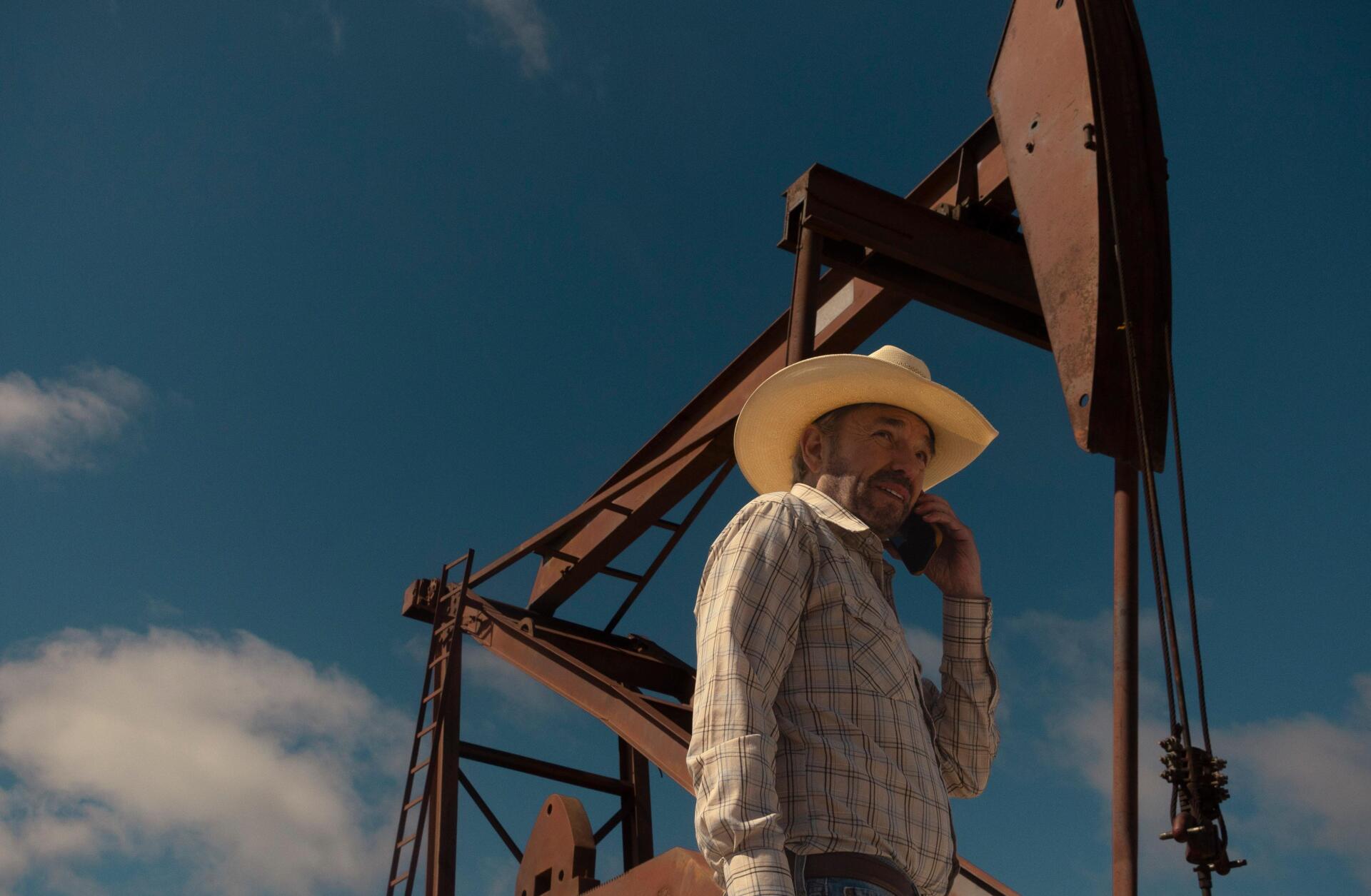 Billy Bob Thornton stands under a clear blue sky, wearing a cowboy hat, with an oil pump jack in the background, showcasing a scene from the TV show Landman.