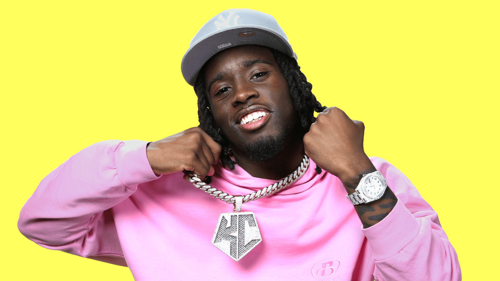 A colorful HD wallpaper featuring streamer Kai Cenat, wearing a pink hoodie and a stylish cap, showcasing a large chain while smiling confidently against a bright yellow background.