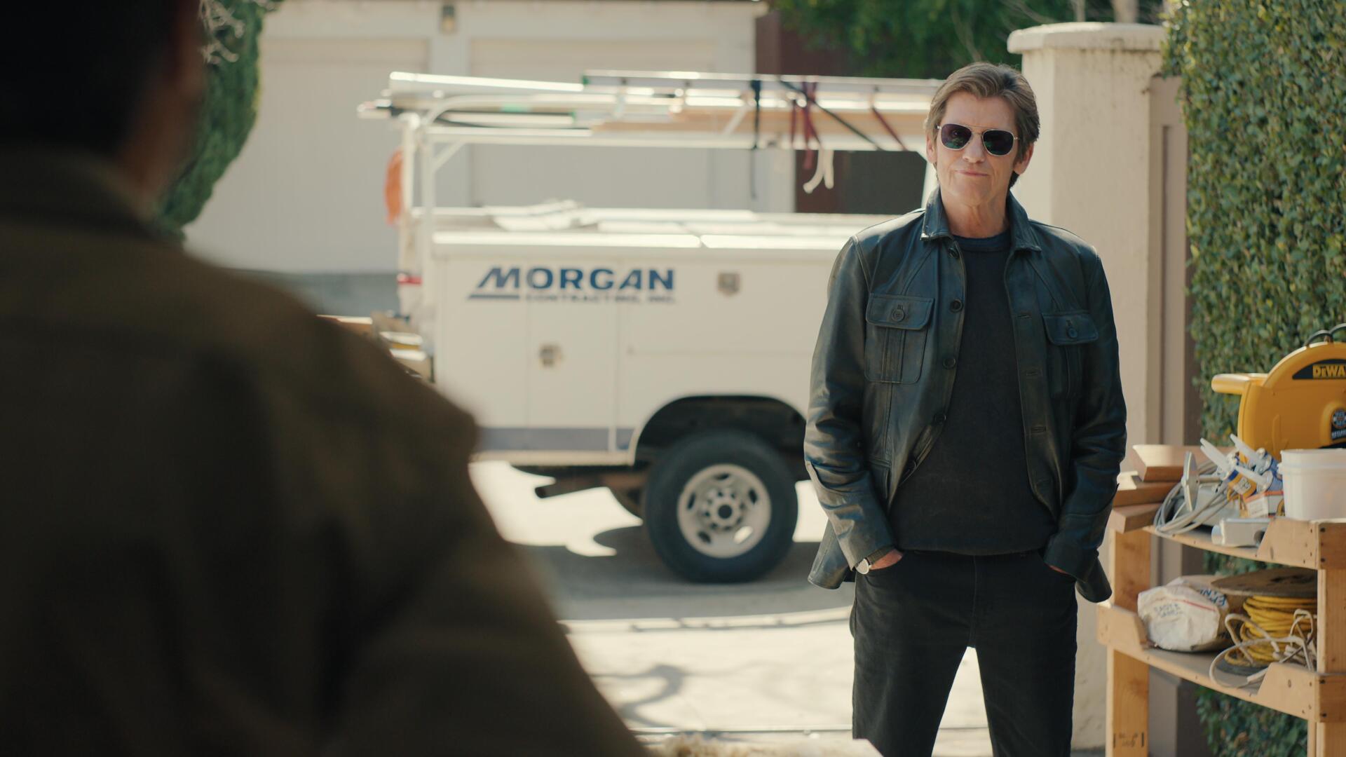 Denis Leary stands confidently in a leather jacket, set against a backdrop featuring a work vehicle, from the TV show No Good Deed (2024). A captivating 4K Ultra HD wallpaper.