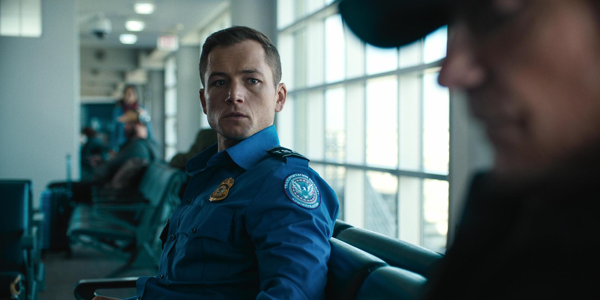 Taron Egerton stars in Carry On (2024), depicted in this HD desktop wallpaper as a focused airport security officer, seated in a modern terminal with large windows.