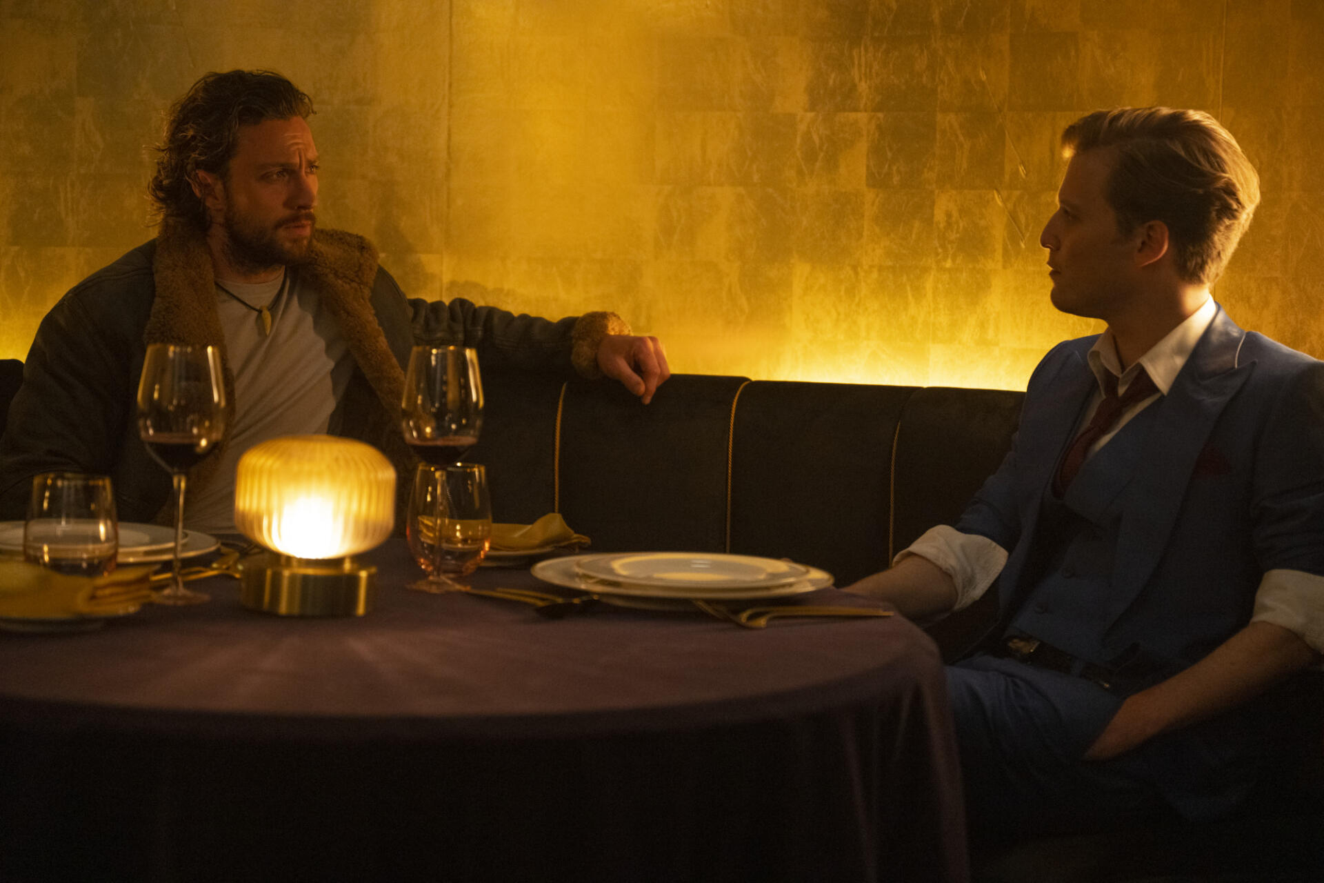 A scene from Kraven the Hunter (2024) featuring Aaron Taylor-Johnson and another character at a table in an elegant setting, with wine glasses and a warm golden ambiance.