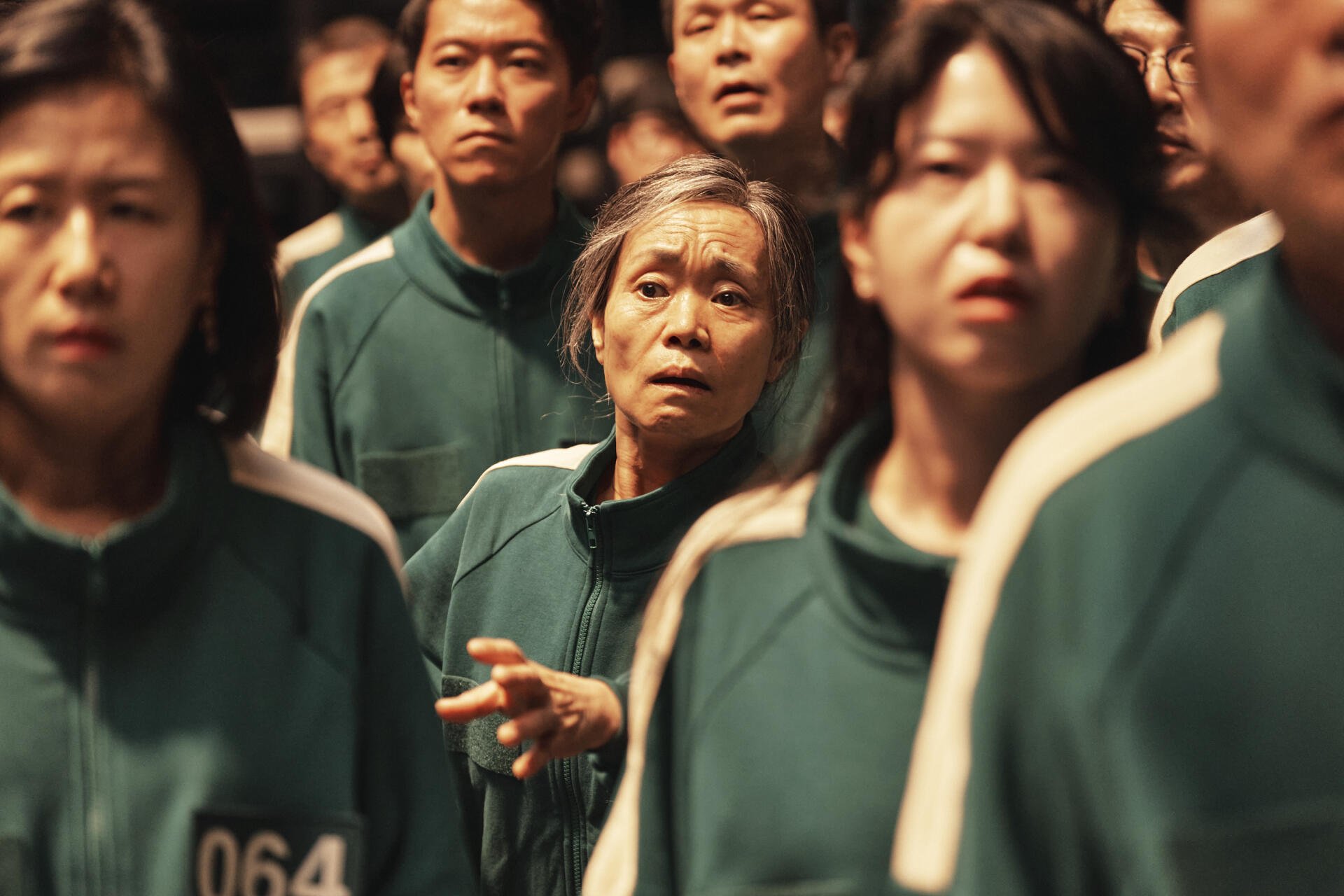 A scene from the TV show Squid Game, featuring a group of participants in green uniforms, showcasing their anxious expressions as they face the challenges of the game.