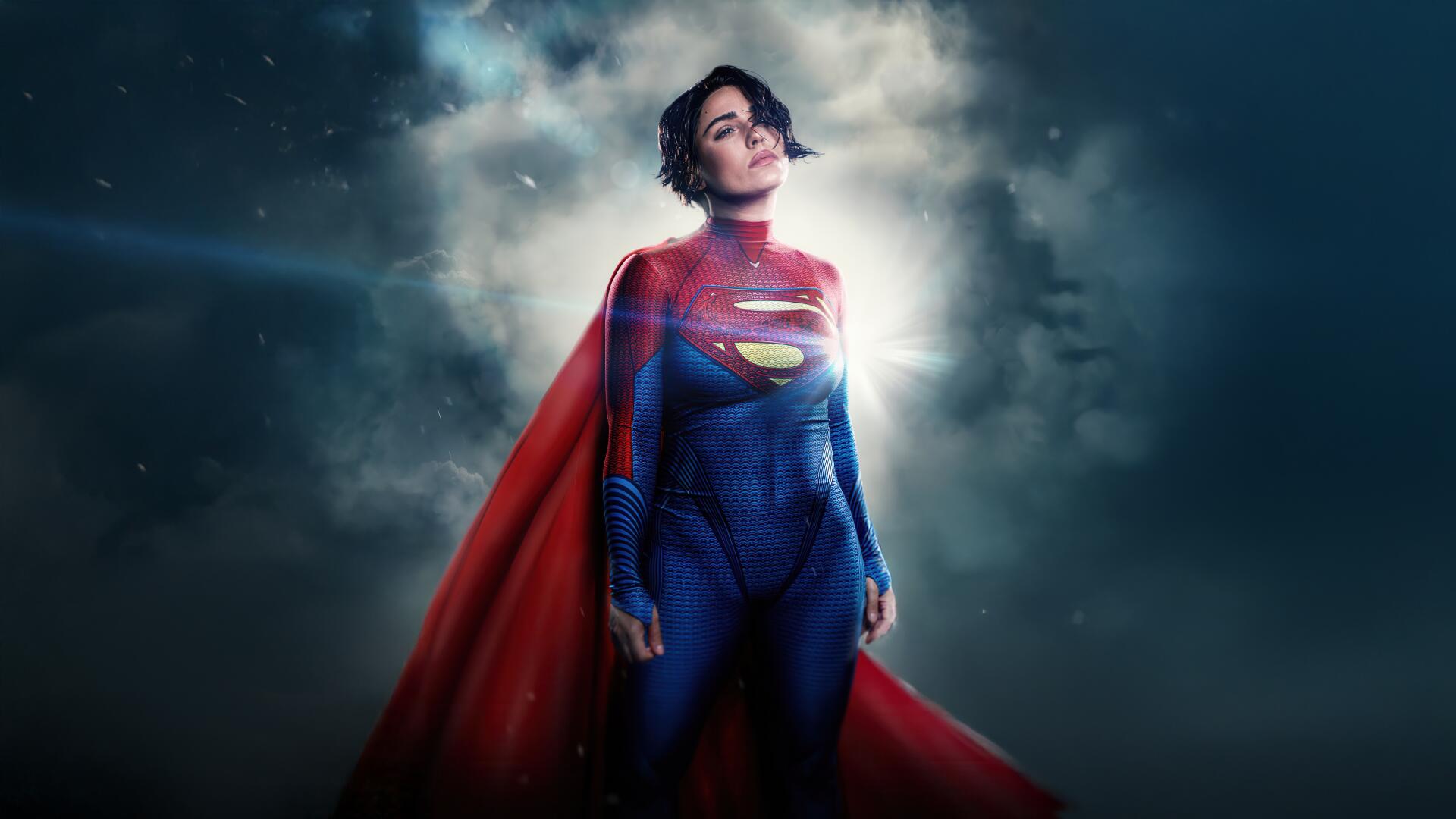 Supergirl portrayal in blue suit and red cape, standing heroically before stormy clouds — 5K Ultra HD PC desktop wallpaper and background.