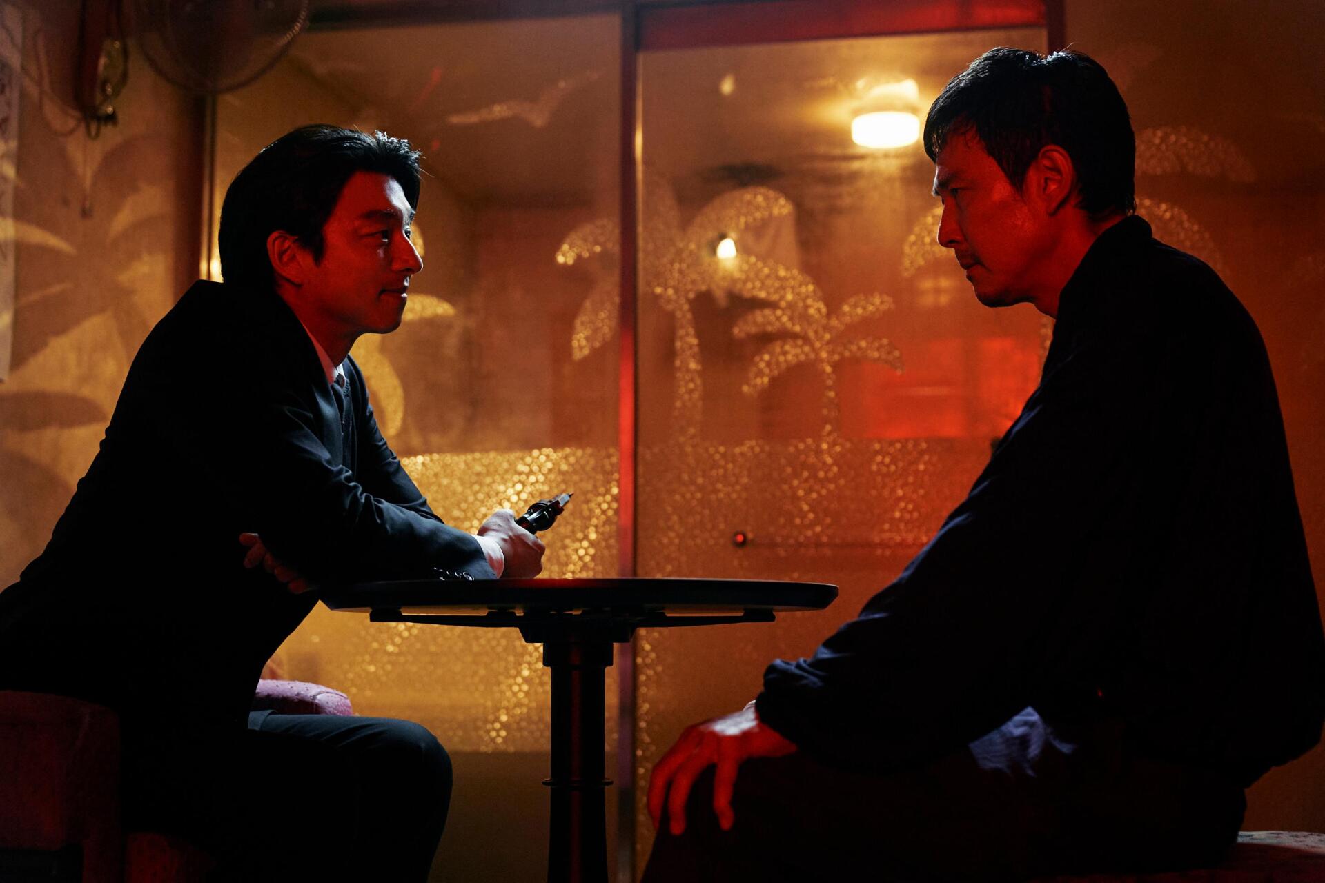 A dramatic scene from the TV show Squid Game, featuring two characters in a tense conversation at a dimly lit table, creating an engaging HD wallpaper for fans.