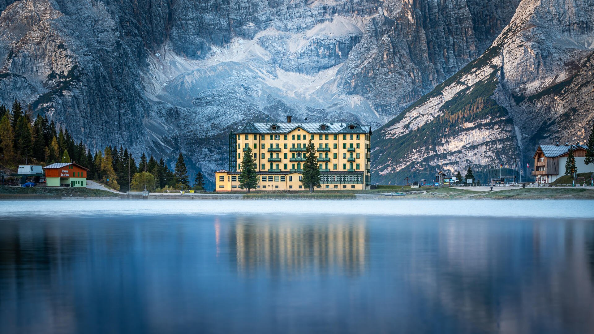 HD PC desktop wallpaper of a serene landscape featuring a large building by a calm sea, set against towering mountainous cliffs under a soft blue sky.