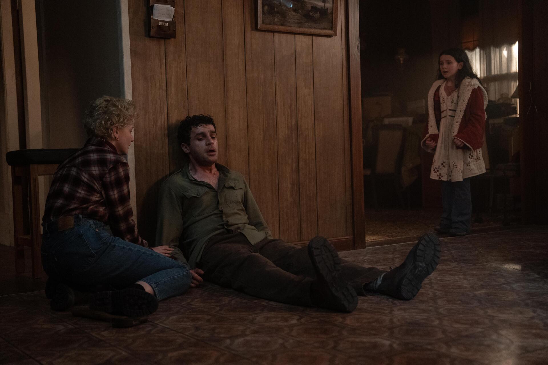 A scene from the movie Wolf Man (2025) depicts a tense moment with a young boy and a man on the floor, while a girl observes from a distance, set against a rustic wooden backdrop.