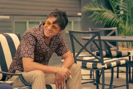 A character from the TV show Tulsa King relaxes outdoors in stylish sunglasses, sporting a patterned shirt, with modern patio furniture and palm plants in the background.