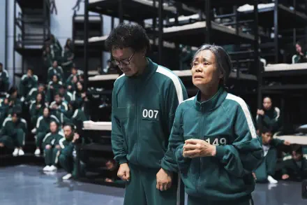 A scene from the TV show Squid Game, featuring characters in green uniforms, expressing tension and emotion in a tense environment filled with other participants.