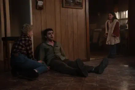 A scene from the movie Wolf Man (2025) depicts a tense moment with a young boy and a man on the floor, while a girl observes from a distance, set against a rustic wooden backdrop.