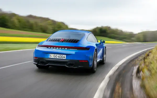 A striking blue Porsche 911 Carrera speeds along a winding road, surrounded by lush greenery and vibrant fields, captured as a high-definition desktop wallpaper.