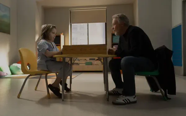 A scene from the TV show Before (2024) featuring Billy Crystal seated across from a young girl at a table, both engaged in a contemplative moment. The setting is bright and simple.