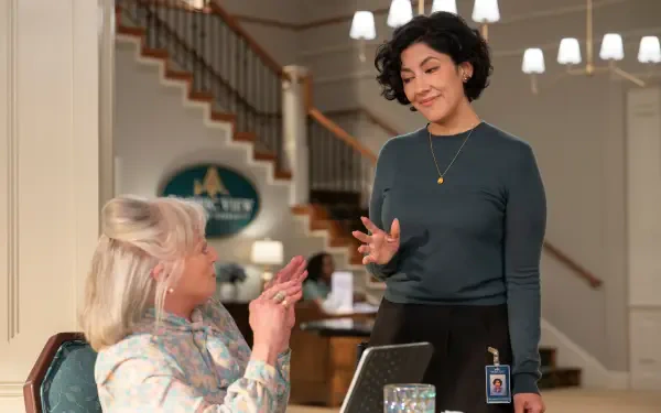 A scene from the TV show A Man on the Inside, featuring two women engaged in conversation. One stands, animated and expressive, while the other listens intently at a table.
