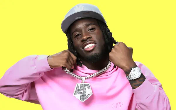 A colorful HD wallpaper featuring streamer Kai Cenat, wearing a pink hoodie and a stylish cap, showcasing a large chain while smiling confidently against a bright yellow background.