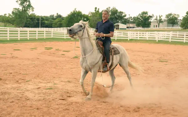 A scene from the TV show Tulsa King, featuring Sylvester Stallone riding a white horse in a dusty arena, set against a backdrop of green trees and a white fence.
