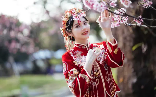 8K Ultra HD PC desktop wallpaper of an Asian woman in ornate red traditional dress smiling among pink cherry blossoms by a pond.