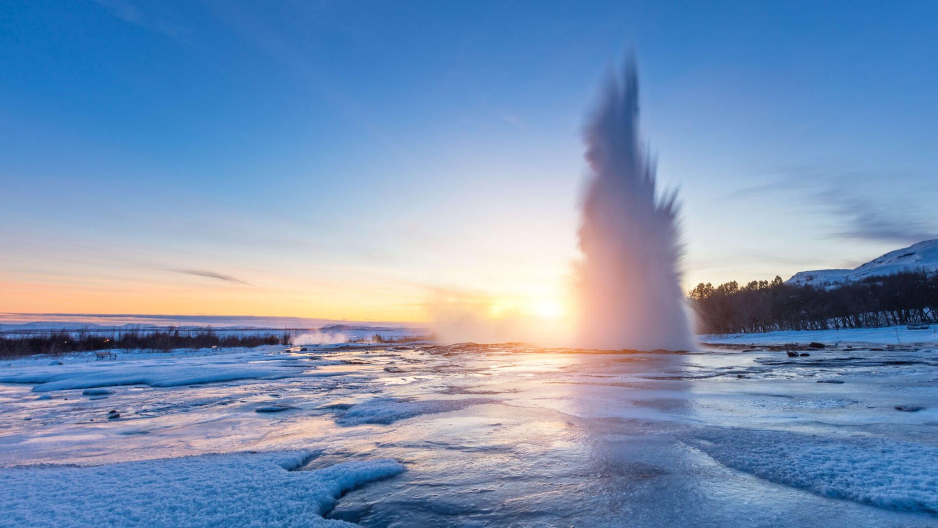 A stunning view of Strokkur Geyser in Iceland, surrounded by snow, erupts against a vibrant sunset sky. This 4K Ultra HD image captures the beauty of nature.
