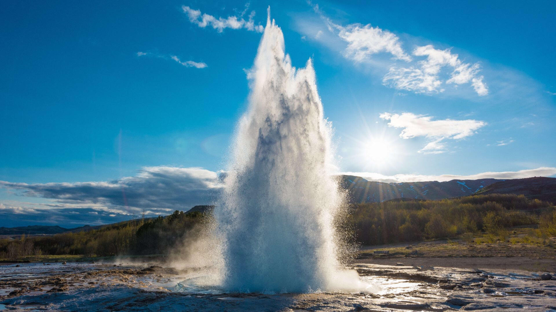 A stunning 4K Ultra HD image of Strokkur Geyser in Iceland, showcasing a powerful water eruption against a bright blue sky and sunlit landscape.