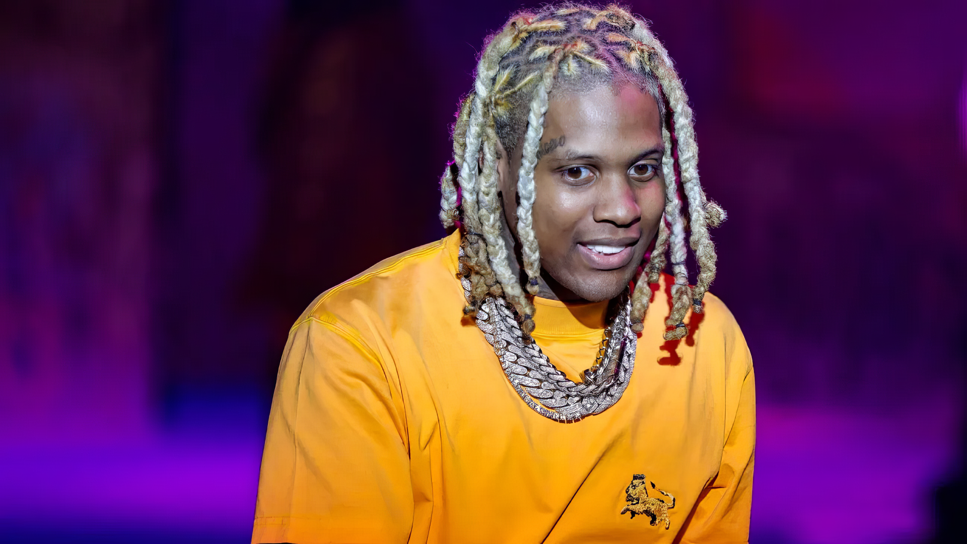 A vibrant HD wallpaper featuring rapper Lil Durk, captured with a playful smile, wearing an eye-catching orange outfit and notable jewelry against a colorful background.