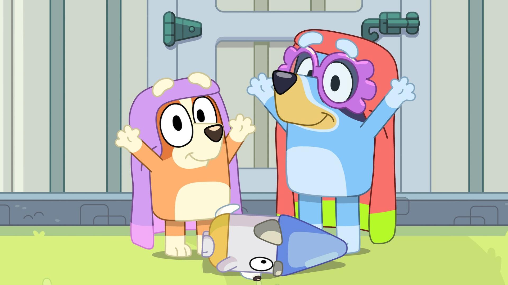 Two playful characters from the TV show Bluey, dressed in colorful capes, joyfully pose with arms raised, while another character lies on the ground, creating a lively scene. HD PC desktop wallpaper.