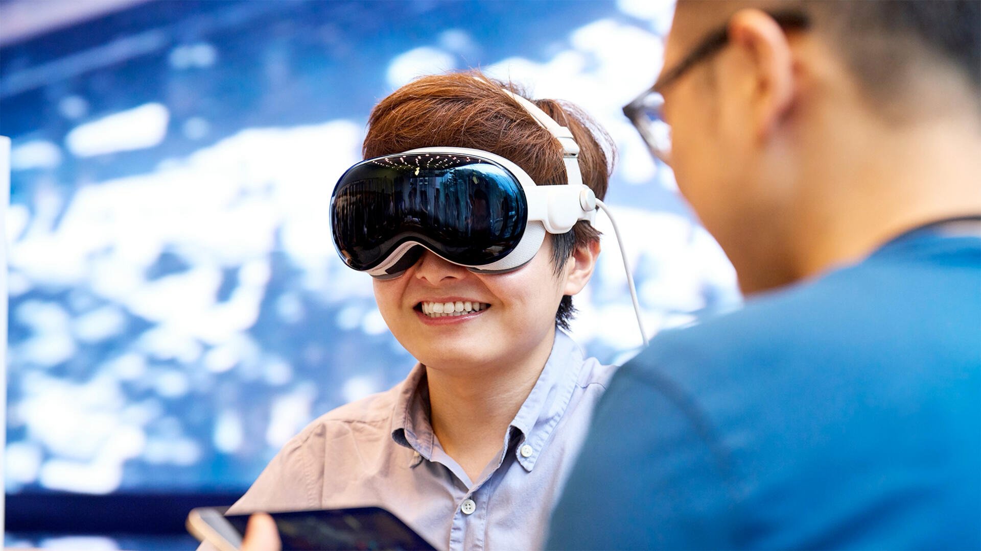 A person wearing Apple Vision Pro goggles smiles while interacting with another individual, showcasing innovative technology by Apple Inc. in a vibrant, high-definition setting.