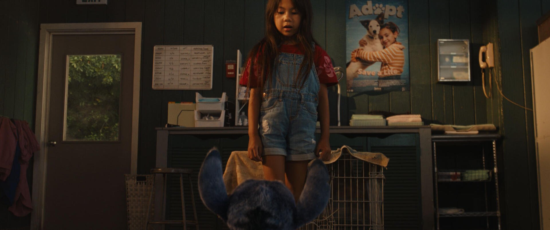 A scene from the movie Lilo & Stitch (2025) shows a young girl in overalls standing over a blue stuffed creature in a room with animal cages and an adoption poster on the wall.