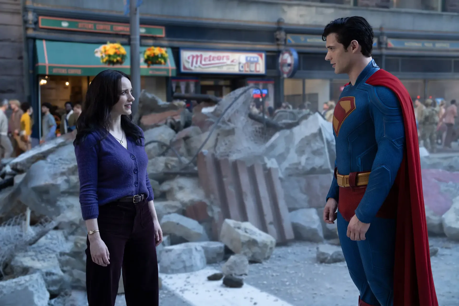 Superman (2025) 2K Quad HD PC desktop wallpaper: Superman in blue suit and red cape stands amid city rubble, facing a woman while onlookers watch from the street.