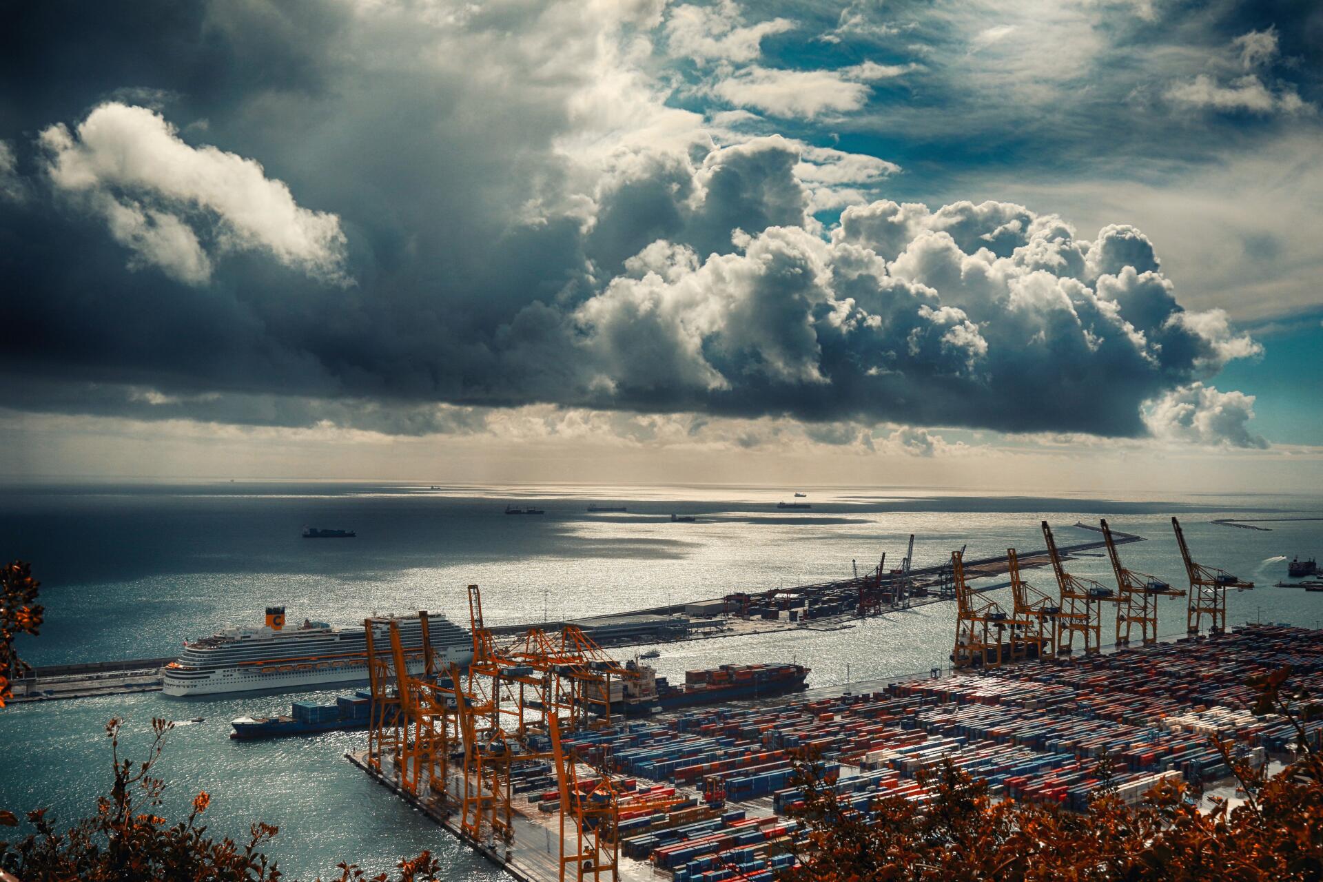 A 4K Ultra HD view of Barcelona's man-made Mediterranean port under dramatic clouds, showcasing shipping containers and cranes along the waterfront.