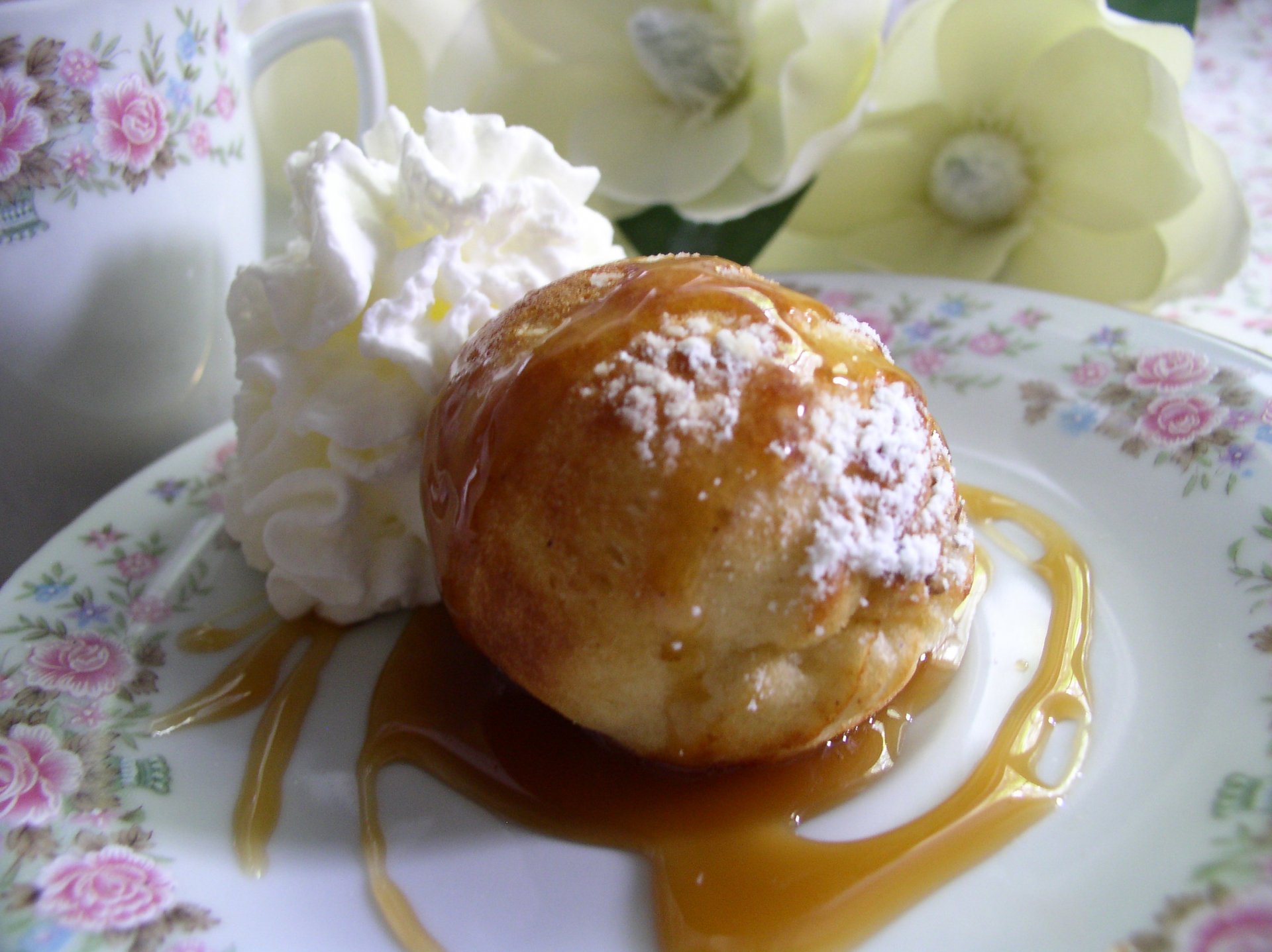 Close-up of a caramel-glazed Danish doughnut dusted with powdered sugar, served with whipped cream on a floral plate.