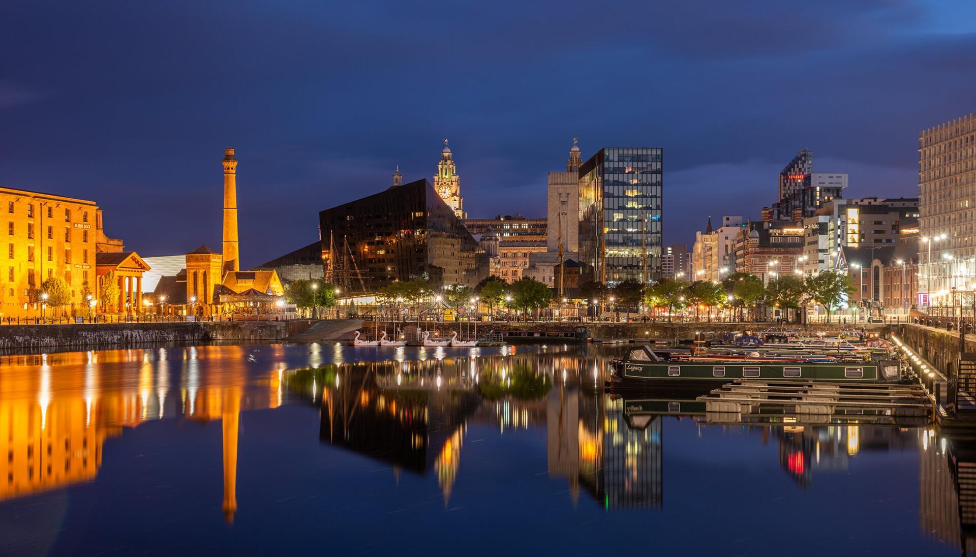 HD PC desktop wallpaper featuring a vibrant night cityscape of Liverpool, with illuminated buildings reflecting on the calm waterfront under a deep blue sky.